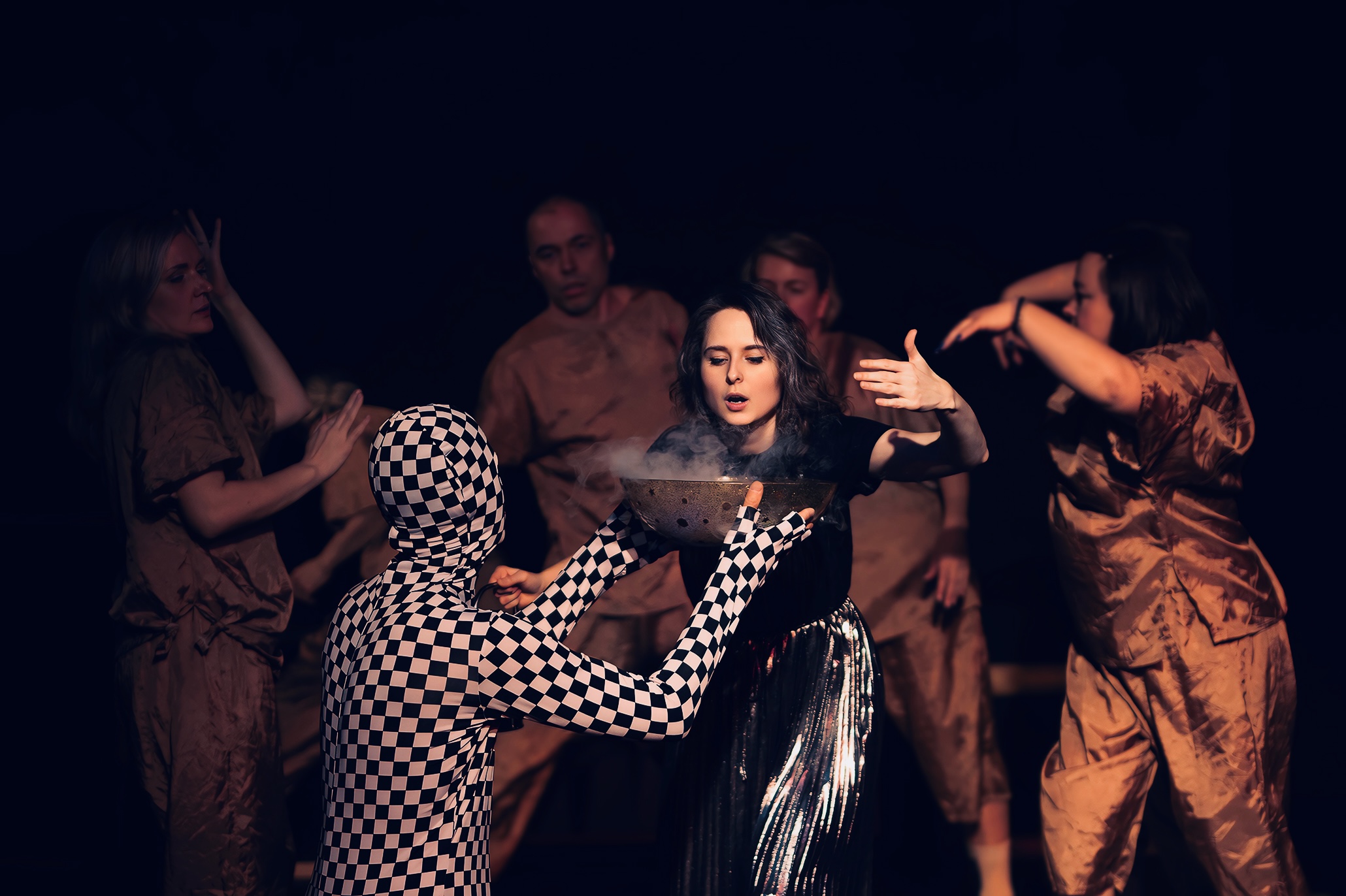 Group of five people on stage, one in checkered costume, others in shiny clothing, performing with expressive gestures.