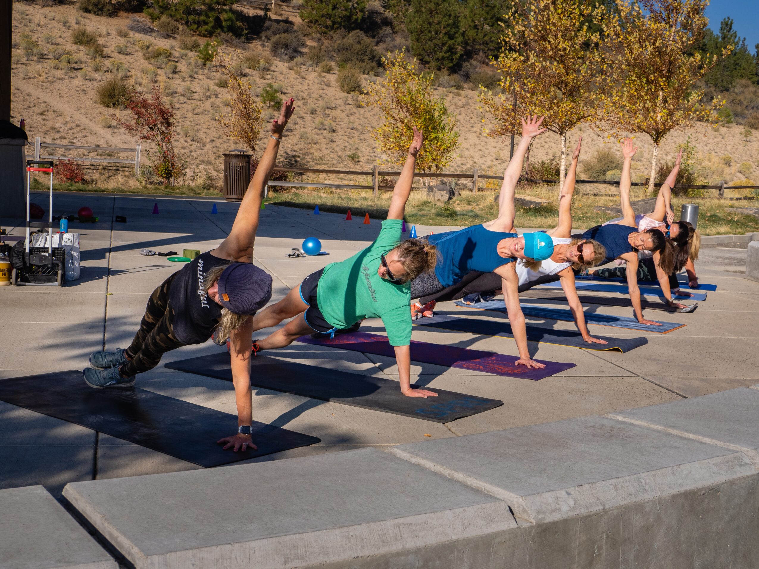 Group of five people doing side plank yoga poses outdoors on mats, with a scenic background of trees and hills.