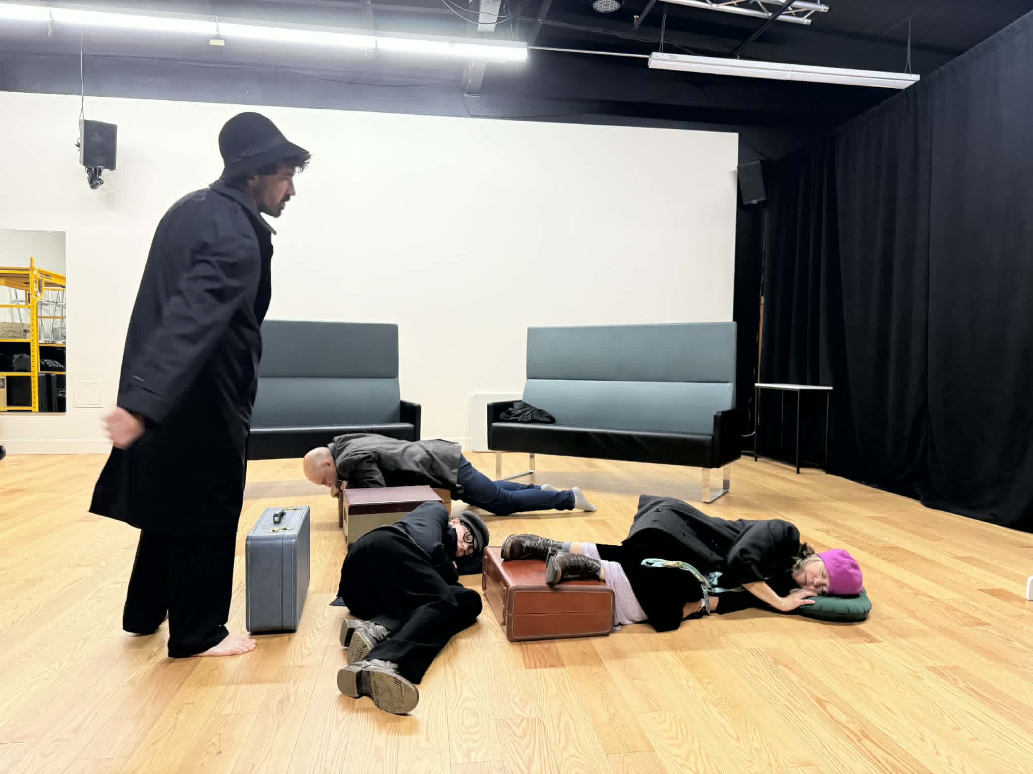 Person standing and three people lying on the floor in a room with wooden floor and black curtains.