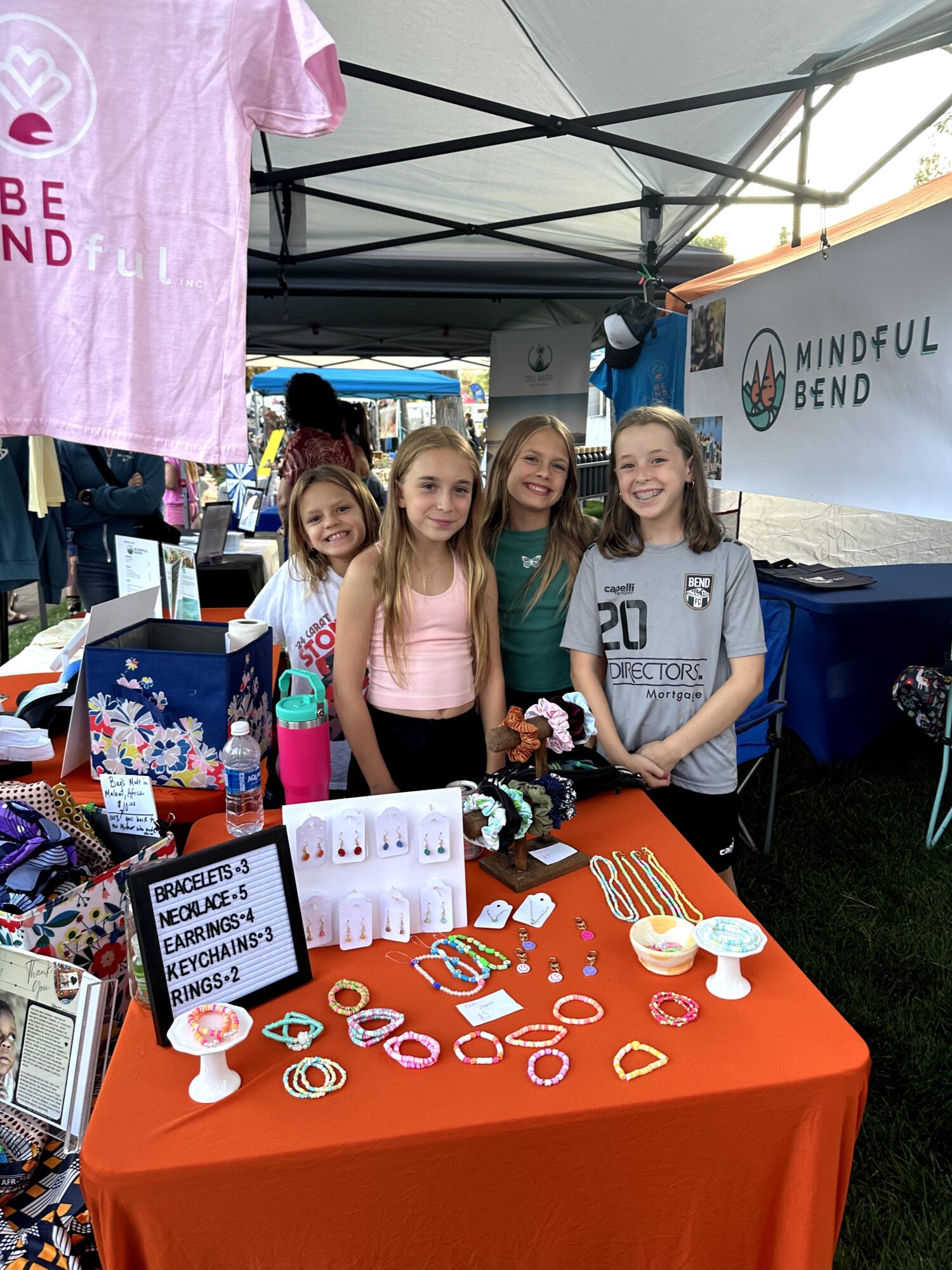 Four children stand behind a table with jewelry and promotional items at an outdoor event under a tent.
