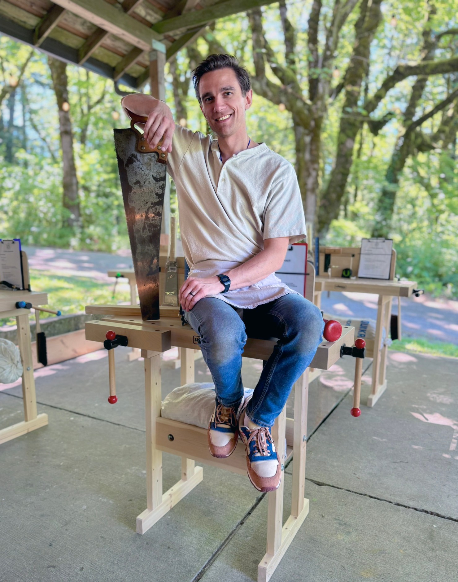 Person sitting on a wooden structure outdoors, smiling, with trees and sunlight in the background.