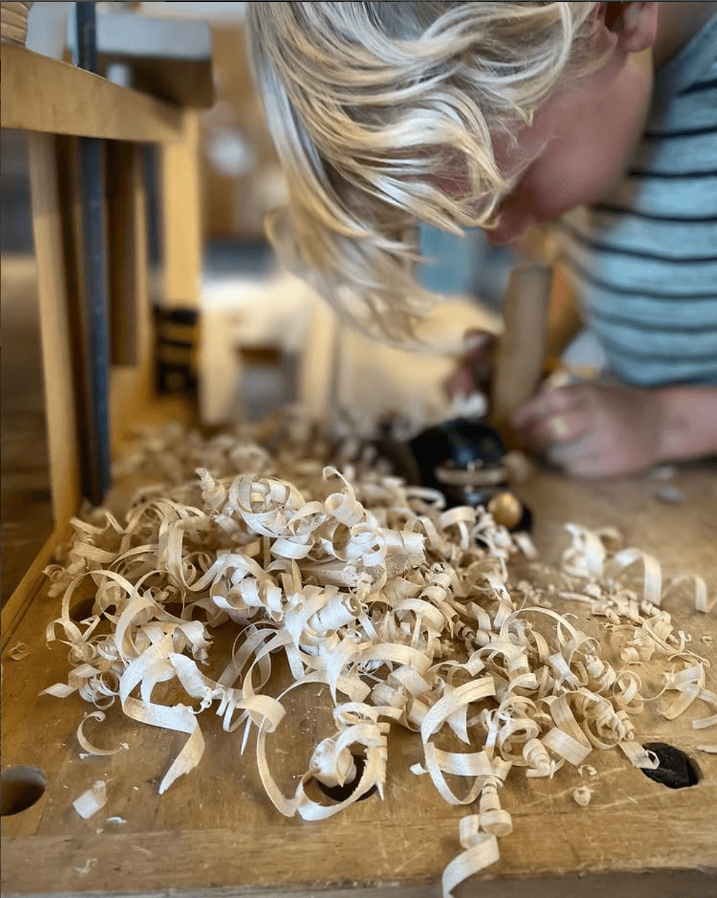 Child with blonde hair leaning over a workbench with wood shavings scattered on it, wearing a striped shirt.