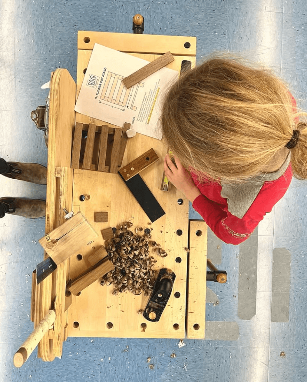 Child with blonde hair in a ponytail, wearing a red shirt, working on a woodworking project at a wooden table.