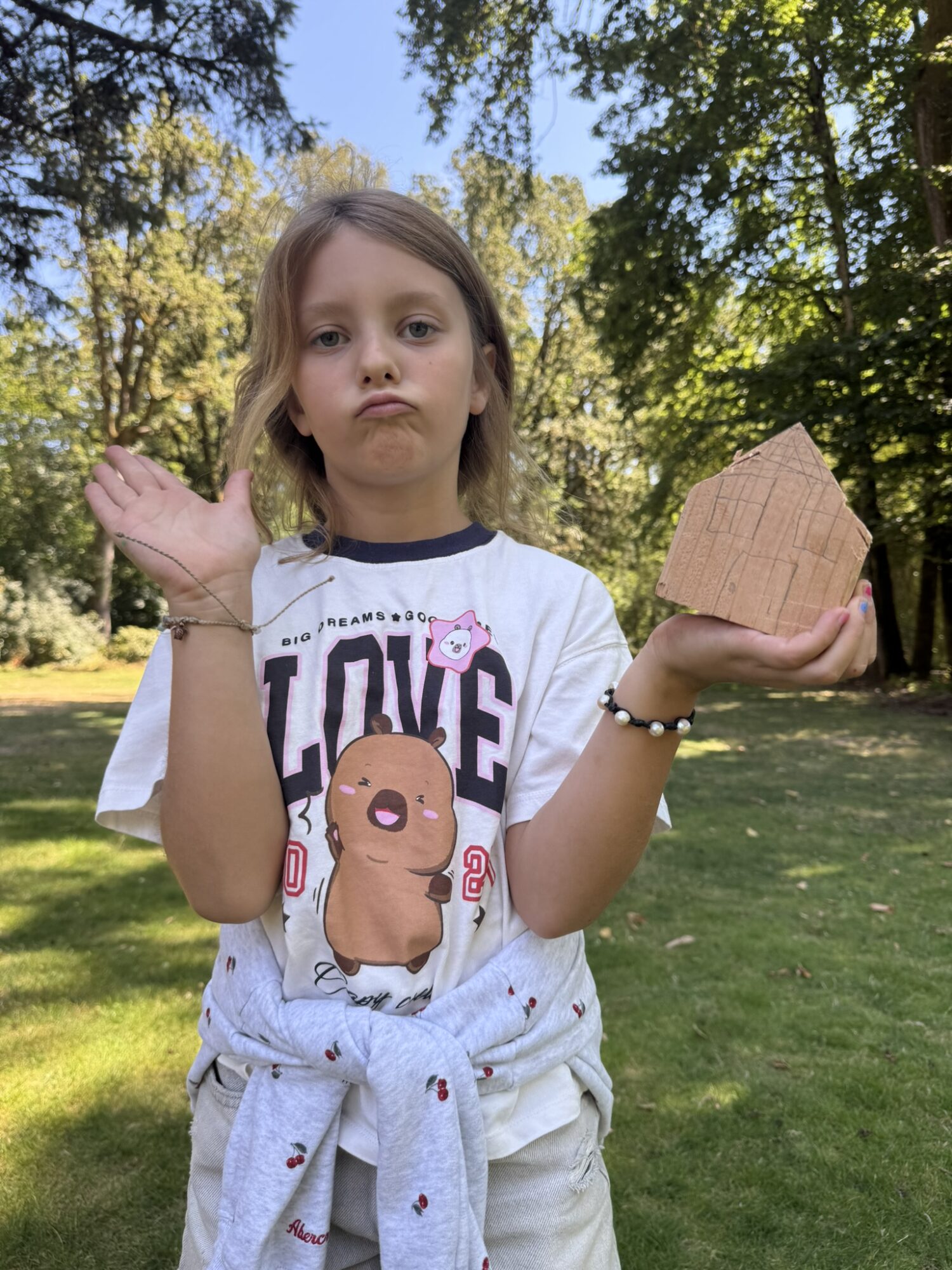 Child standing outdoors holding a paper house in one hand and raising the other hand, trees and sky in background.