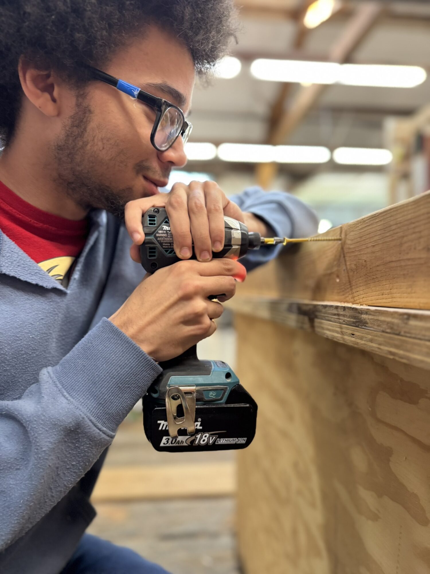 Young man with glasses using a cordless drill on a wooden surface in a workshop.