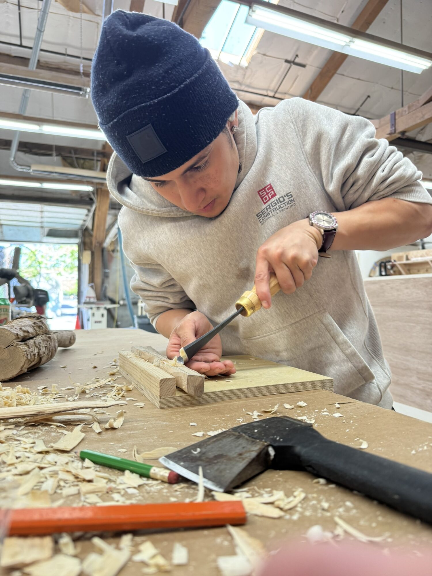 Young person wearing a beanie and sweatshirt using a chisel on a woodworking project in a workshop.