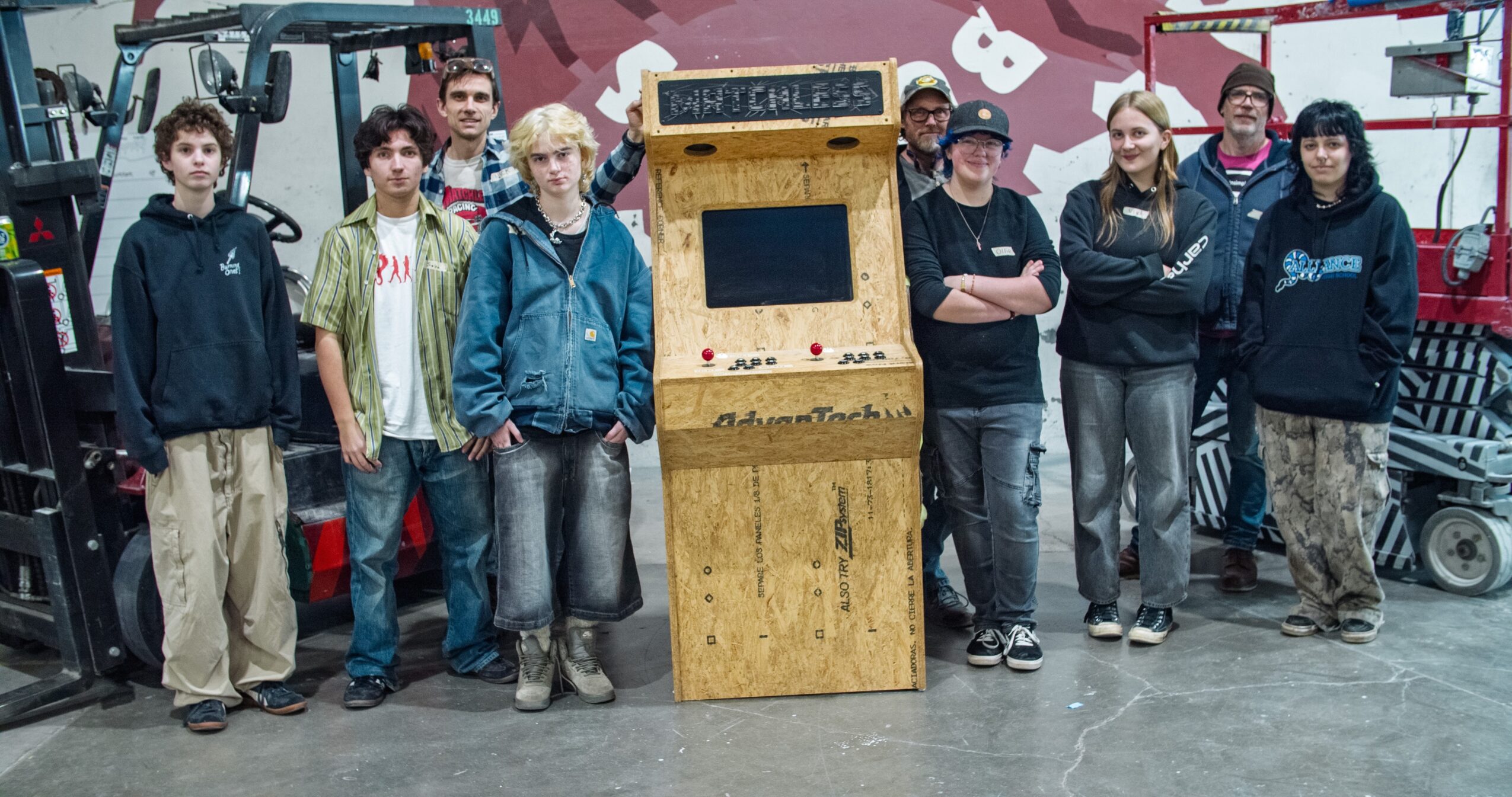 Group of eight people standing in front of a wooden arcade game in an industrial setting.