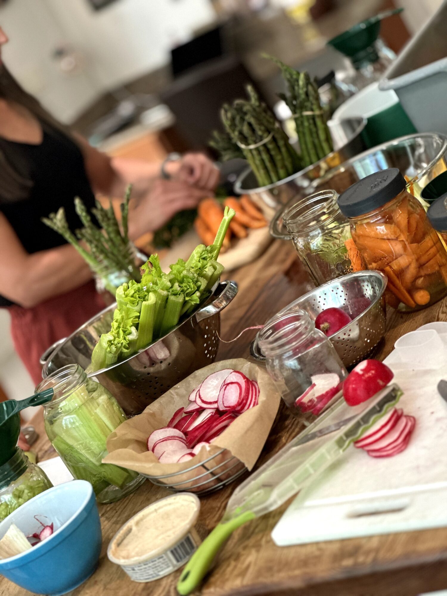 Fresh vegetables, including radishes, celery, and cucumbers, arranged on a wooden table with jars of pickles and a person preparing food.
