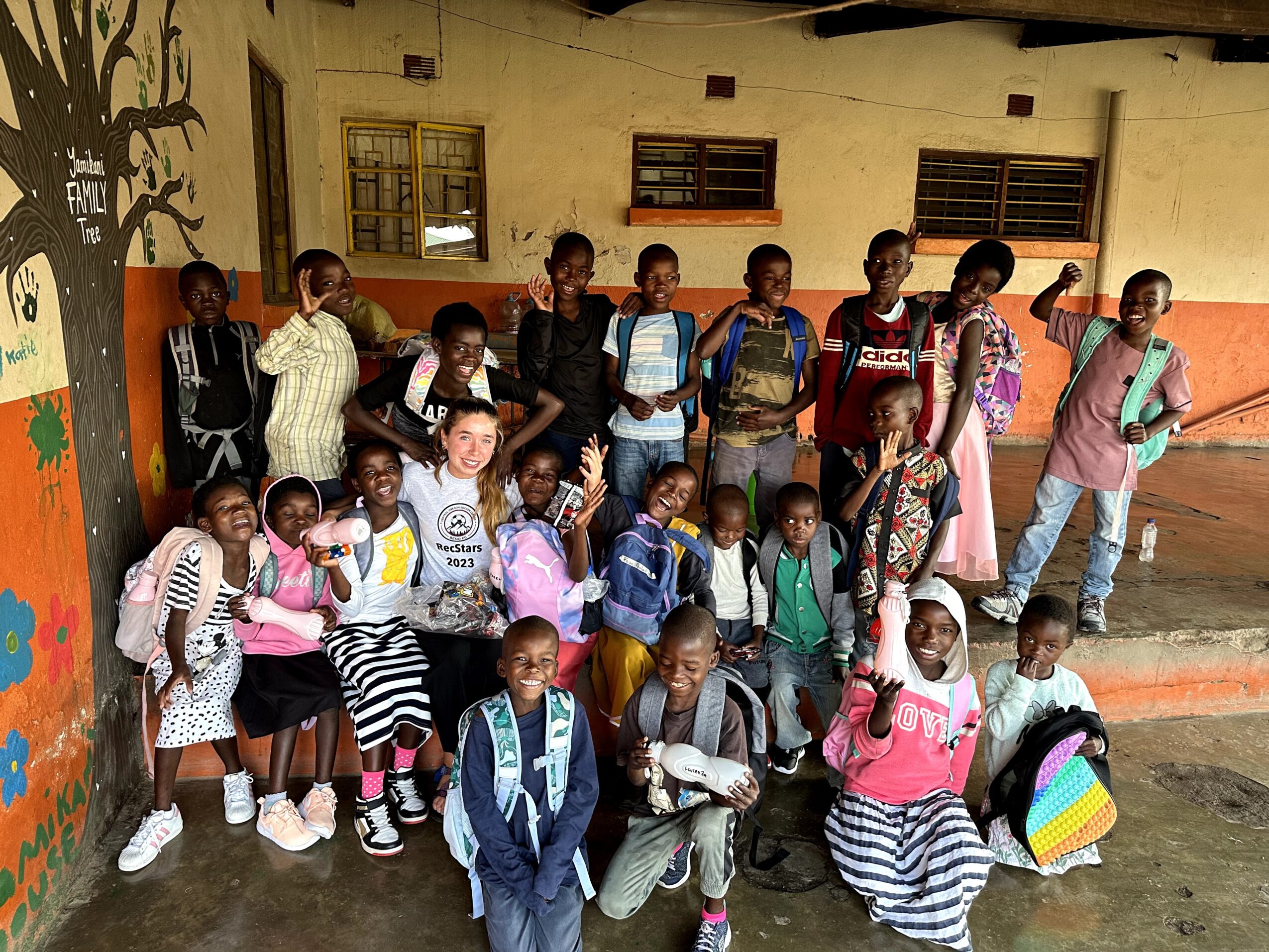 Group of children and teenagers standing and sitting outside a building with windows and a door, some holding backpacks.