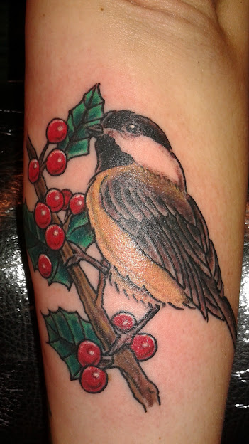 Colorful bird perched on a branch with holly leaves and red berries, detailed tattoo on skin.