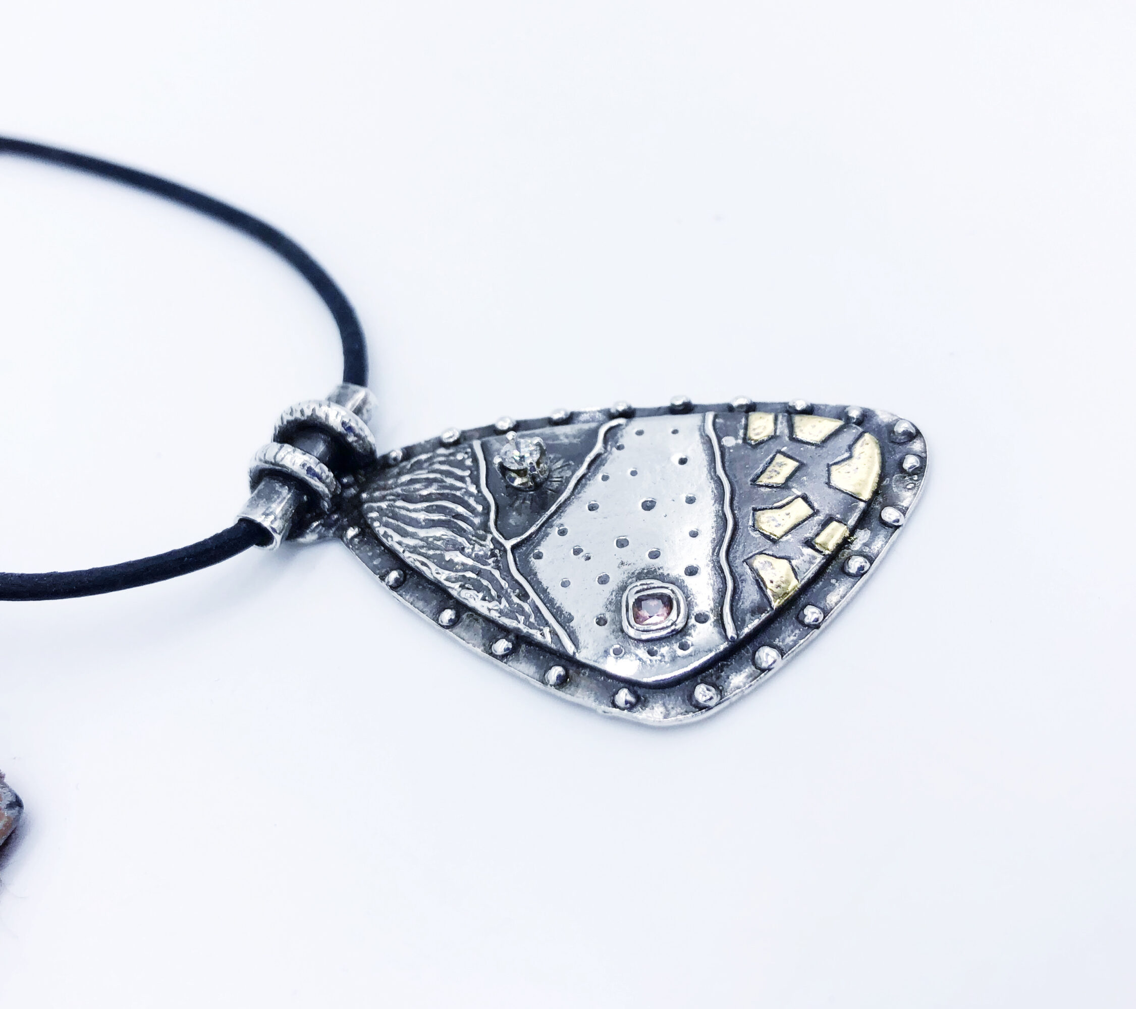 Pendant necklace with a large, irregularly shaped metallic pendant on a black cord, featuring textured and patterned surfaces.