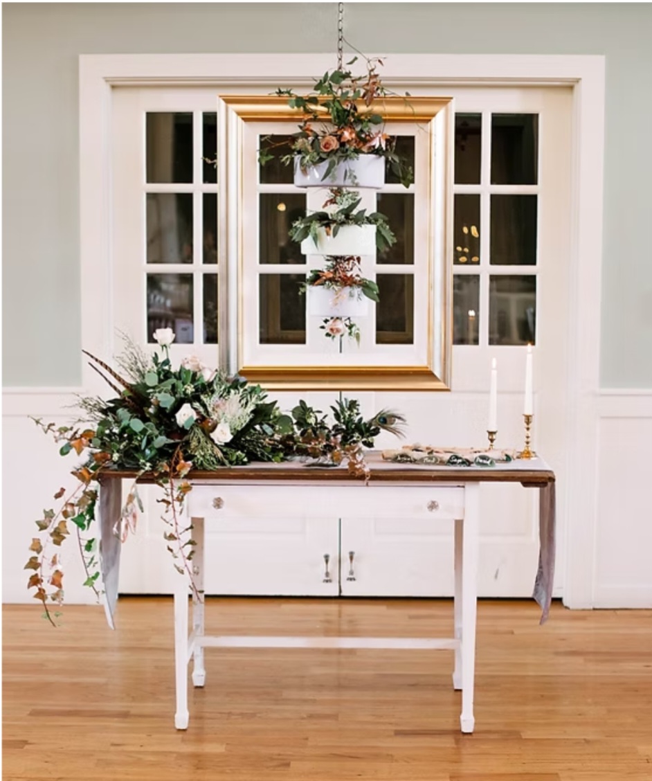 Decorative table with floral arrangement and candles in front of a window with hanging floral display, wooden floor, and white walls.