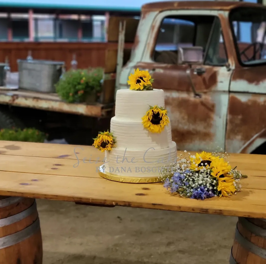 White cake decorated with yellow sunflowers and purple flowers on a wooden table outdoors, with an old rusty truck in the background.