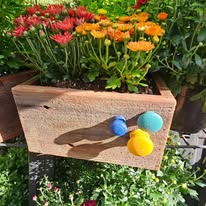 Wooden planter box with pink, yellow, and orange flowers, with colorful balls attached, outdoors in sunlight.