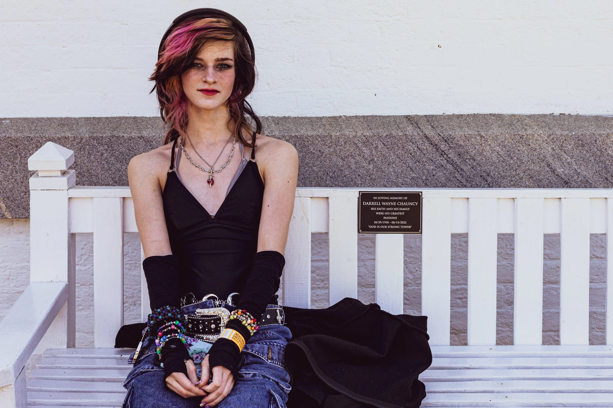 Young woman with dark hair and makeup sitting on a white bench against a white wall, wearing a black dress and accessories.