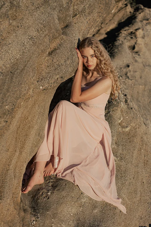 Young woman with curly hair sitting on rocks, wearing a light pink dress, with a rocky background.