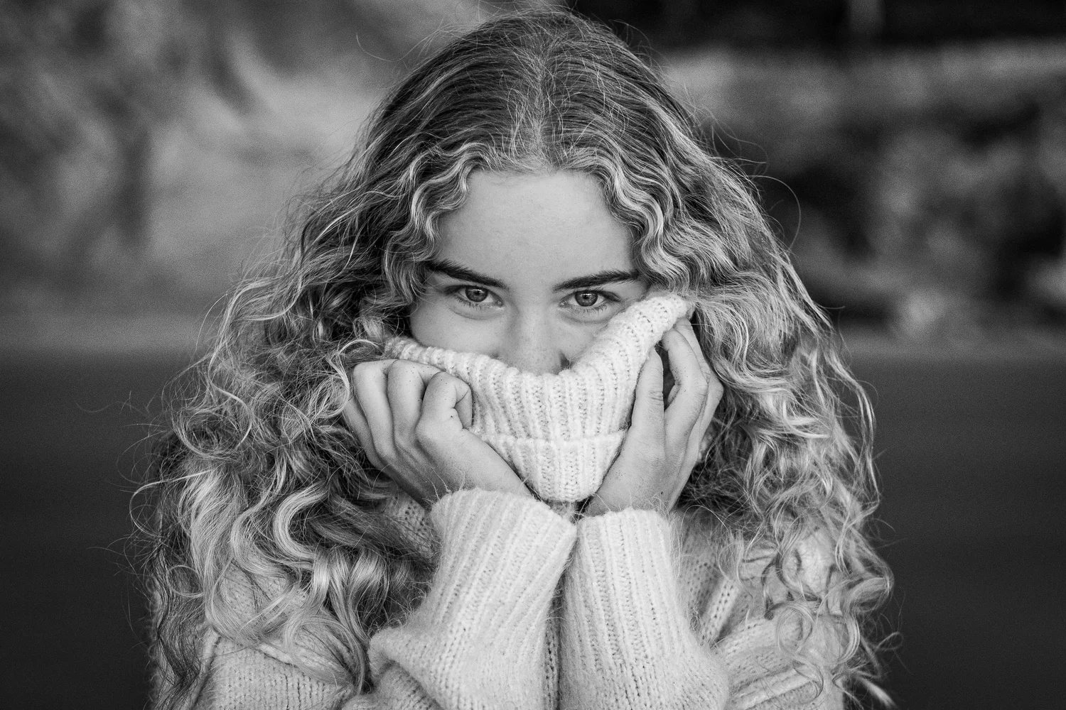 Young woman with curly hair covering her face with a sweater, eyes visible, outdoors, black and white.
