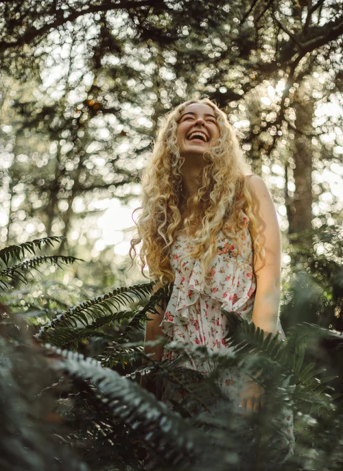 Woman with long curly hair smiling and looking up in a forest setting surrounded by trees and ferns.