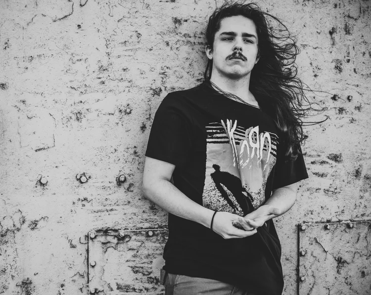 Young person with long hair standing against a textured wall, wearing a graphic t-shirt and holding an object.