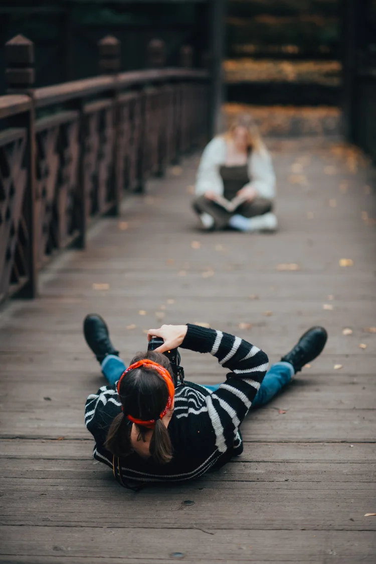 Person lying on wooden bridge with head turned, holding a phone, and another person sitting in the background.