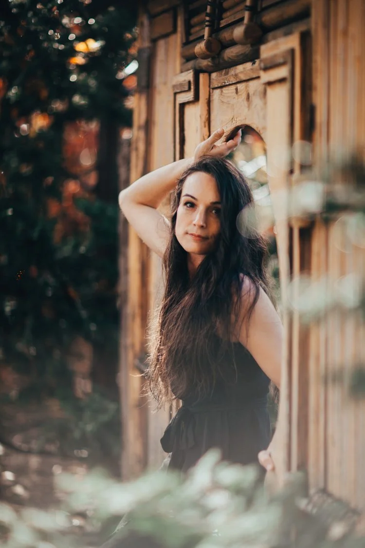Young woman with long dark hair standing outdoors near a wooden structure, looking at the camera with her hand on her head.