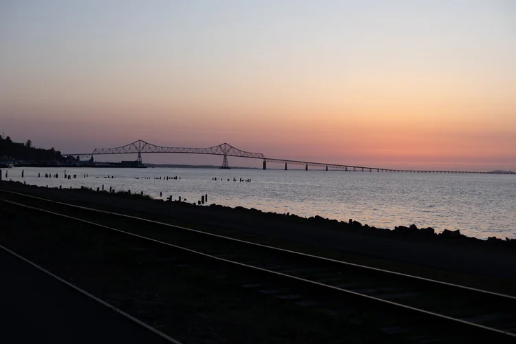 Sunset over water with a bridge in the distance, dark shoreline, and train tracks in foreground.