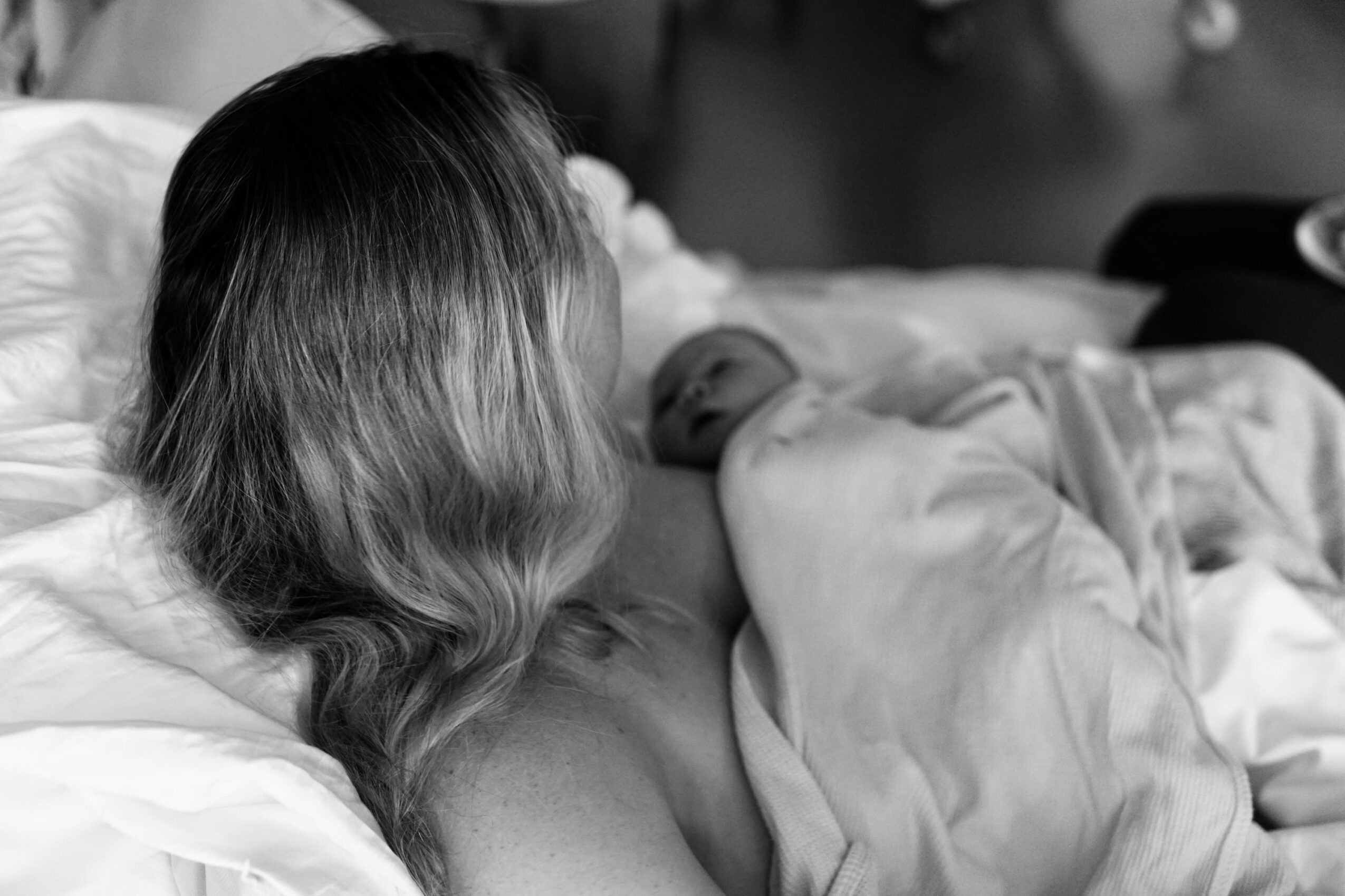 Woman holding a newborn baby on a bed, with the woman’s face turned away from the camera, in black and white.