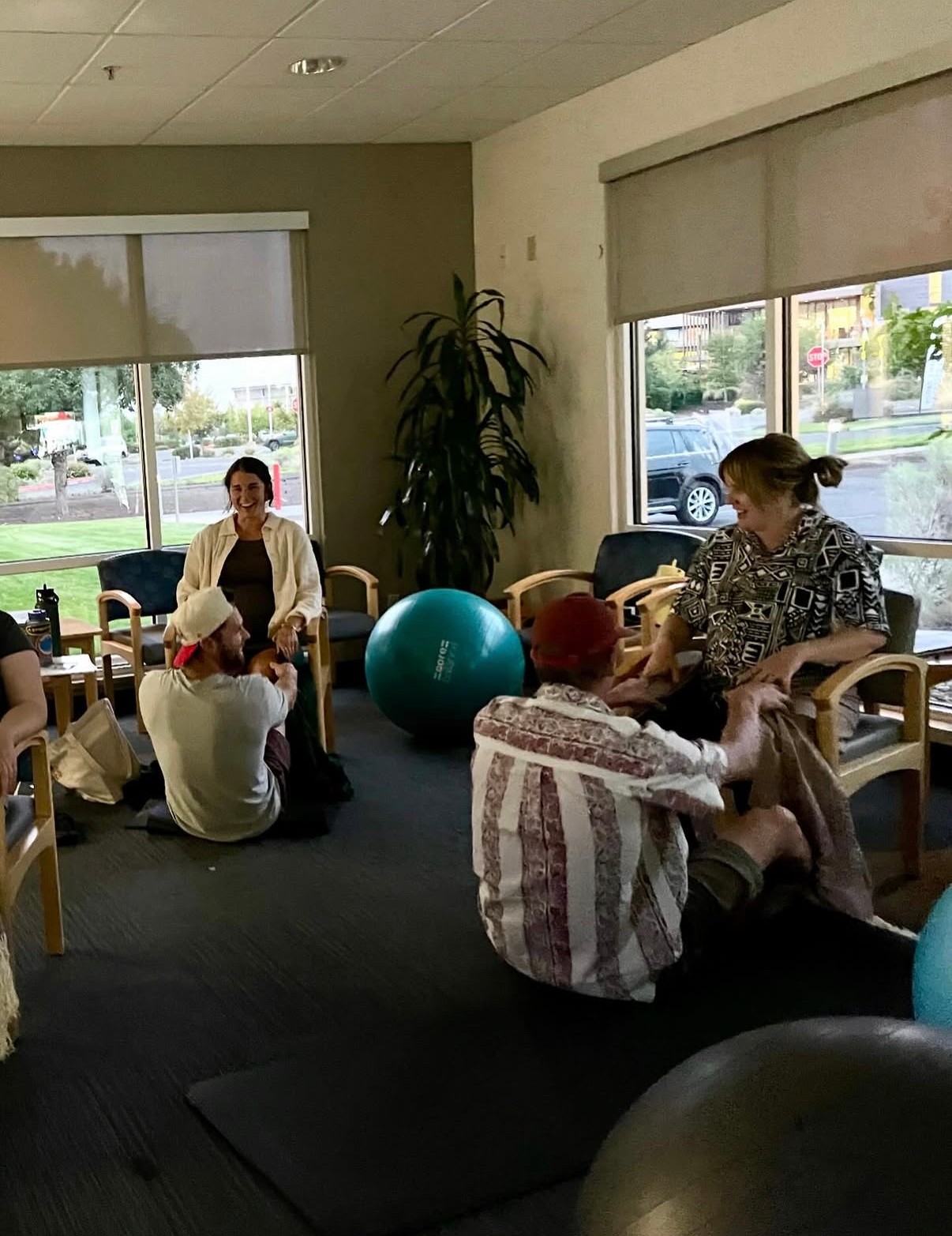 Group of people sitting and talking in a room with large windows, a potted plant, and exercise balls.