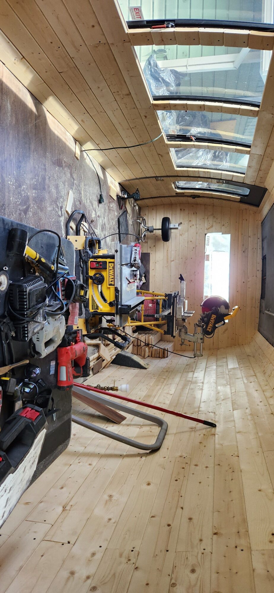 Workshop with tools, equipment, and a ladder inside a wooden-paneled room with windows, viewed from the side.