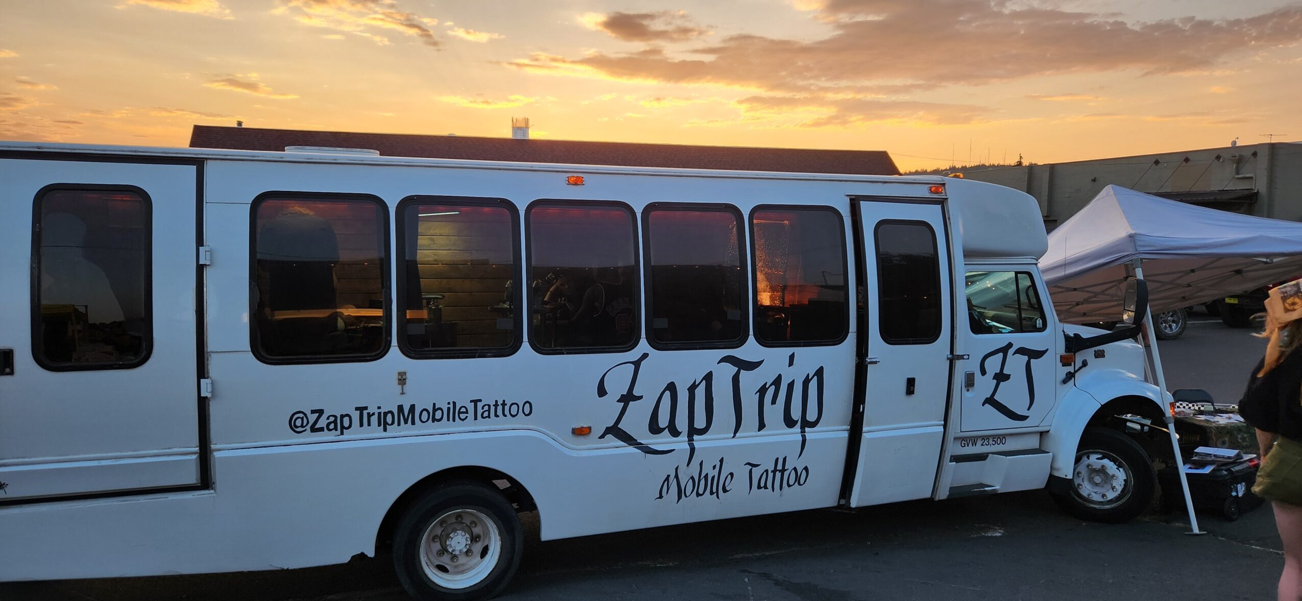 White mobile tattoo bus parked outdoors during sunset, with a tent nearby and a person standing beside it.