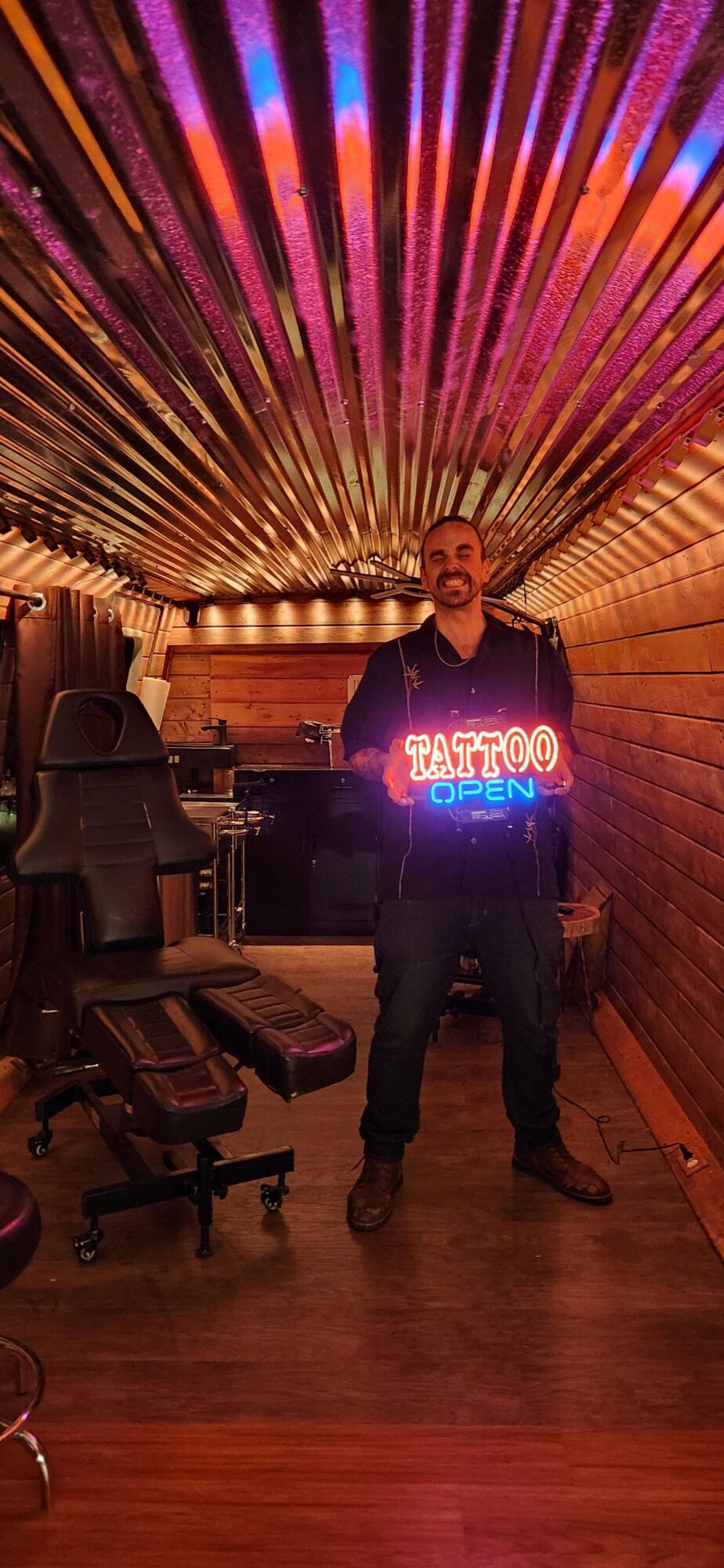 Person standing in a room with a neon sign, wooden walls, and chairs, facing the camera.