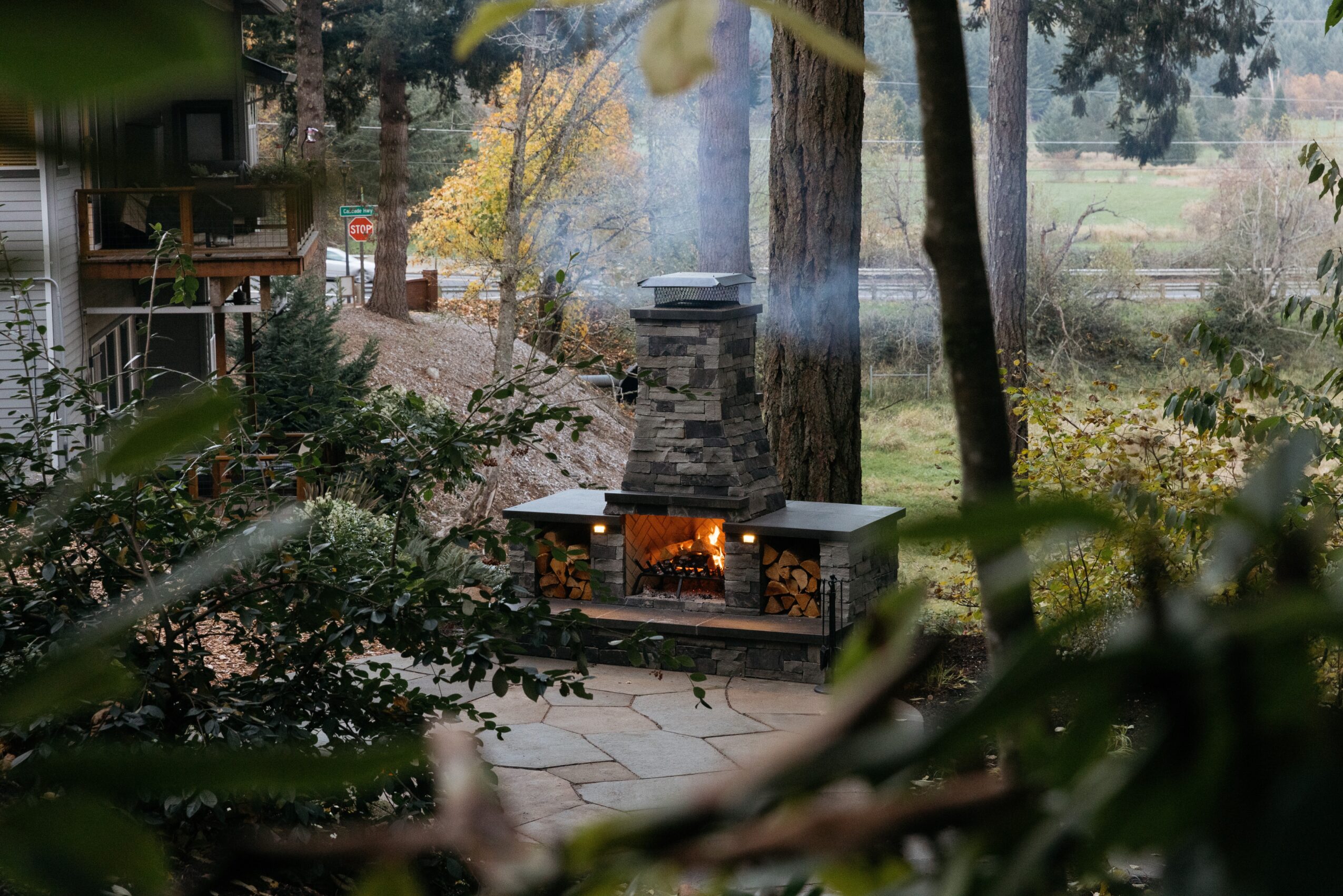 Outdoor fireplace with a chimney, surrounded by trees and plants, in a garden setting, with a stone patio.