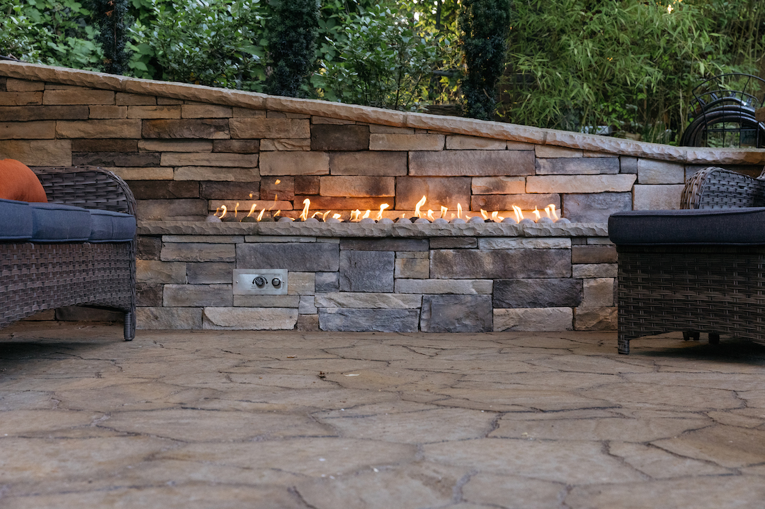 Outdoor patio with a stone wall and a linear fire feature, two wicker chairs, and trees in the background.