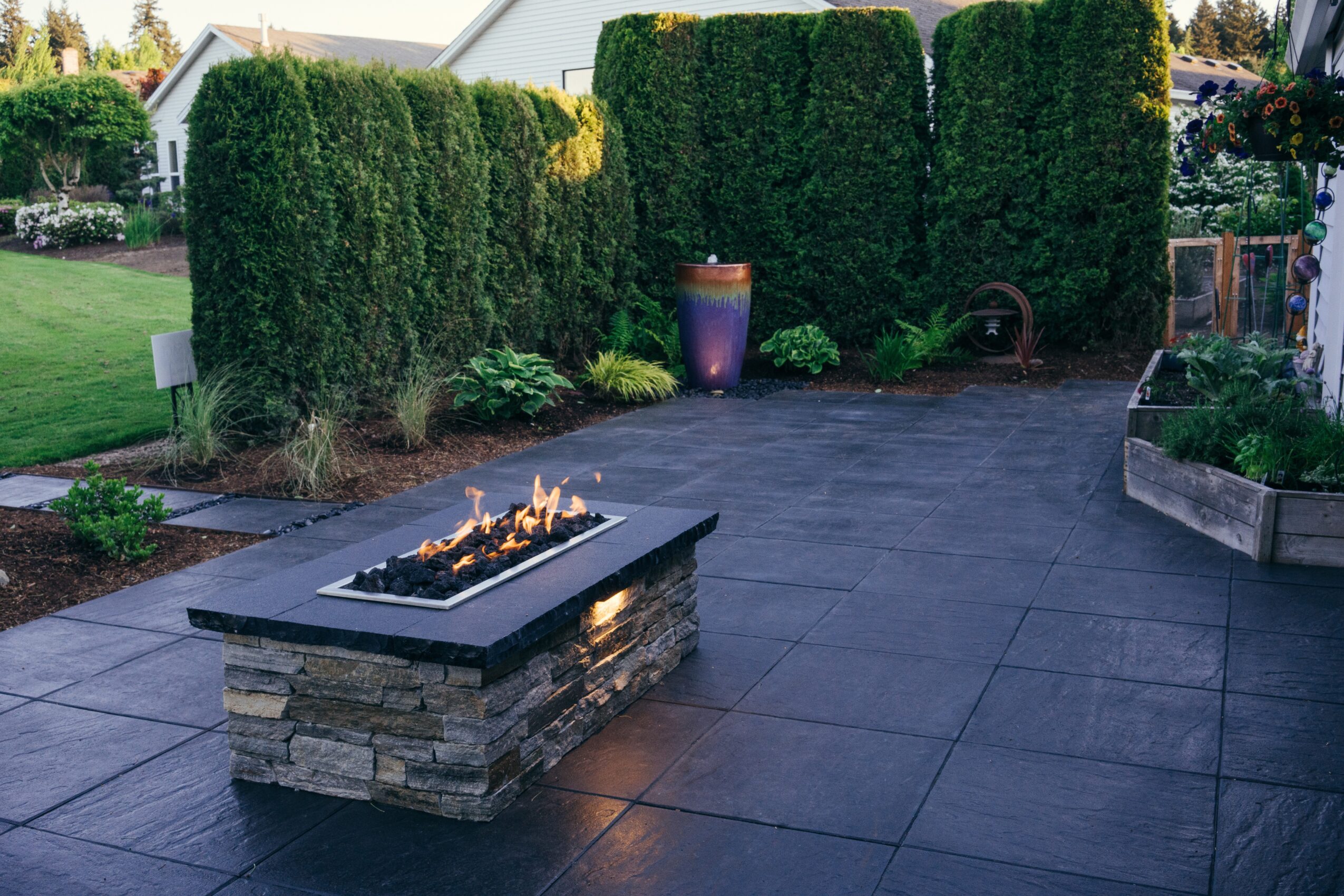 Outdoor patio with a fire feature, tall green hedges, potted plants, and a paved surface.