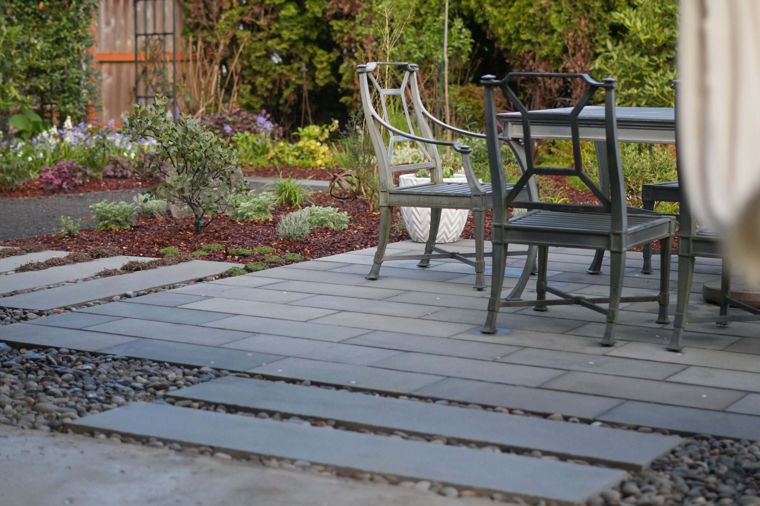 Outdoor patio with chairs and table, garden with plants and trees in background, paved stone pathway, and gravel area.