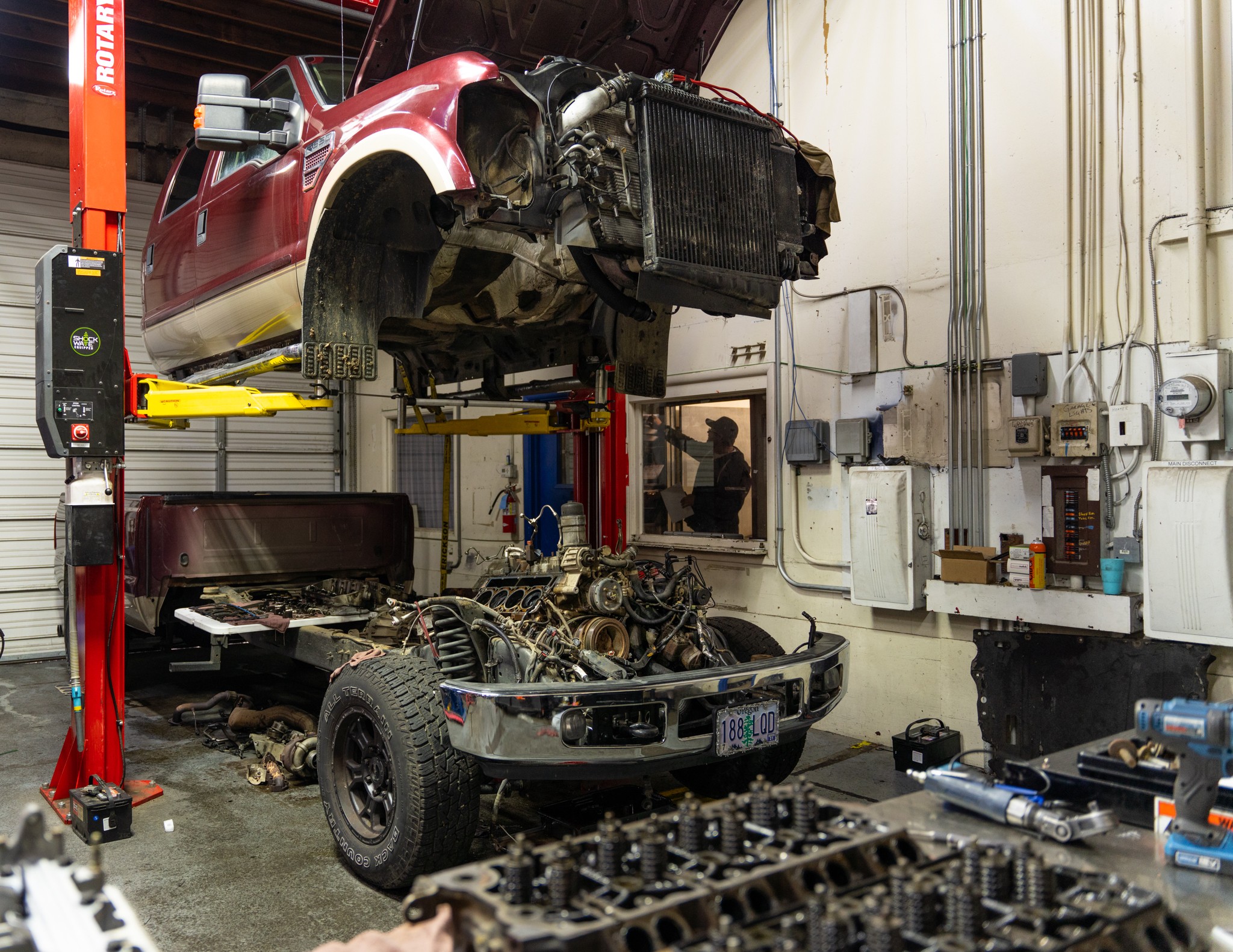 Red pickup truck lifted on a hydraulic lift in a garage, with the front removed and engine exposed, tools nearby.