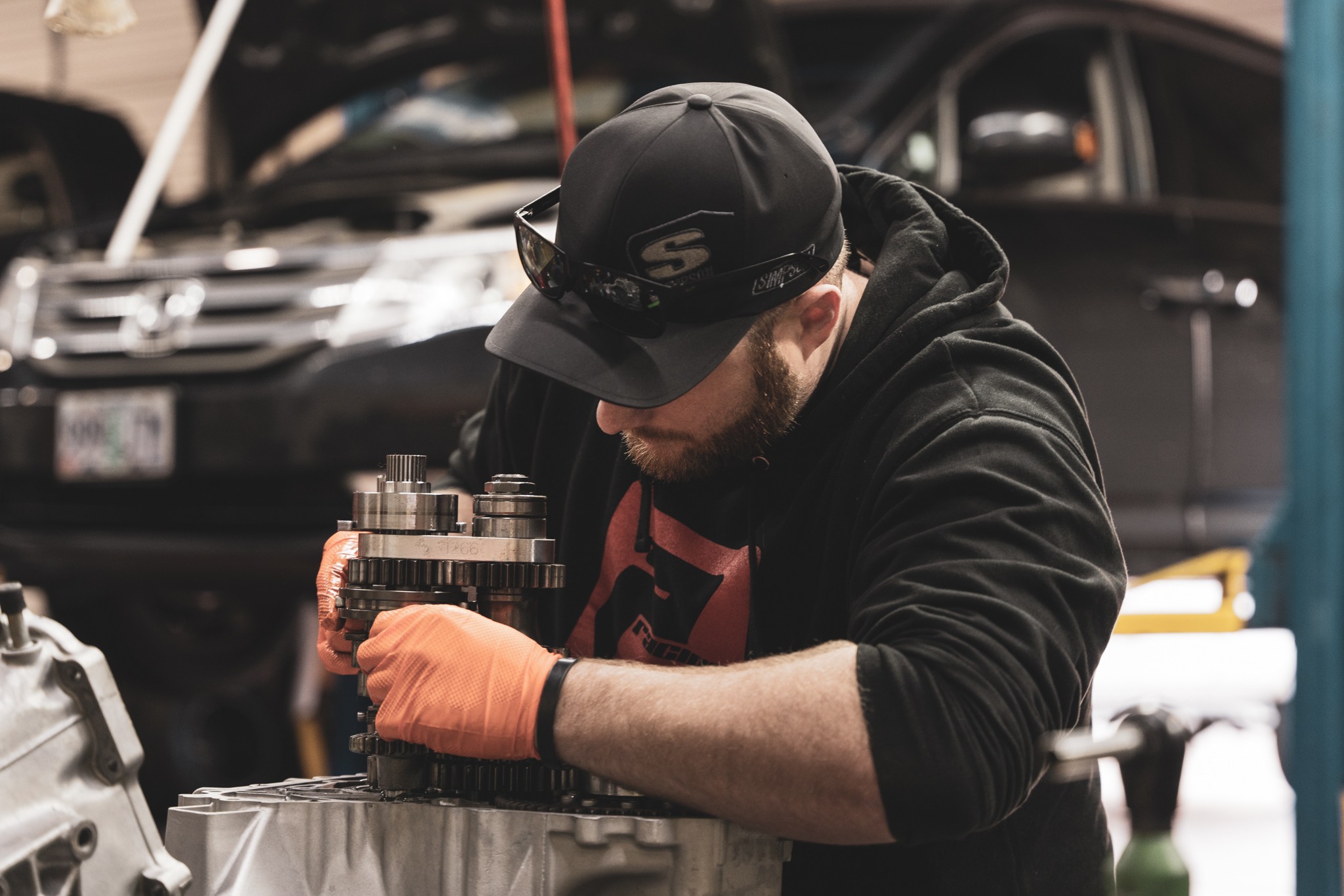 Man working on a mechanical component outdoors, wearing a black cap, glasses, black hoodie, and orange gloves.
