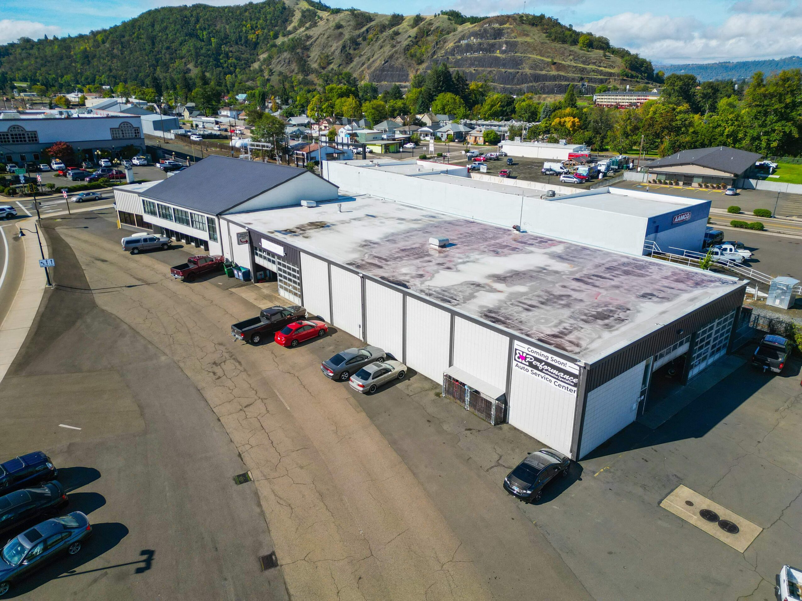 Large warehouse building with multiple loading docks, surrounded by parked cars and a parking lot, with a hill and trees in the background.