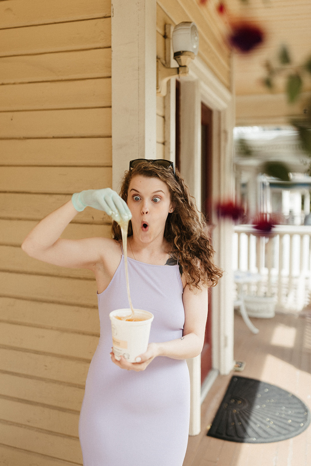 Young woman with curly hair wearing a light purple dress, holding a bowl and pouring liquid, standing outside a house.