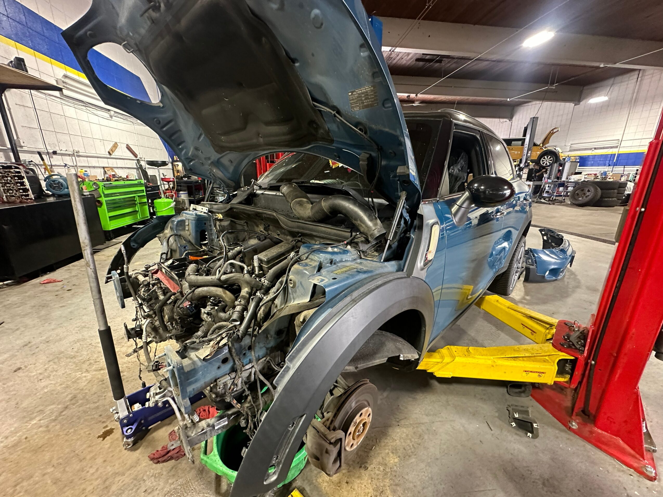 Blue vehicle with open hood in a garage, lifted on a yellow hydraulic lift, engine removed, tools and equipment around.