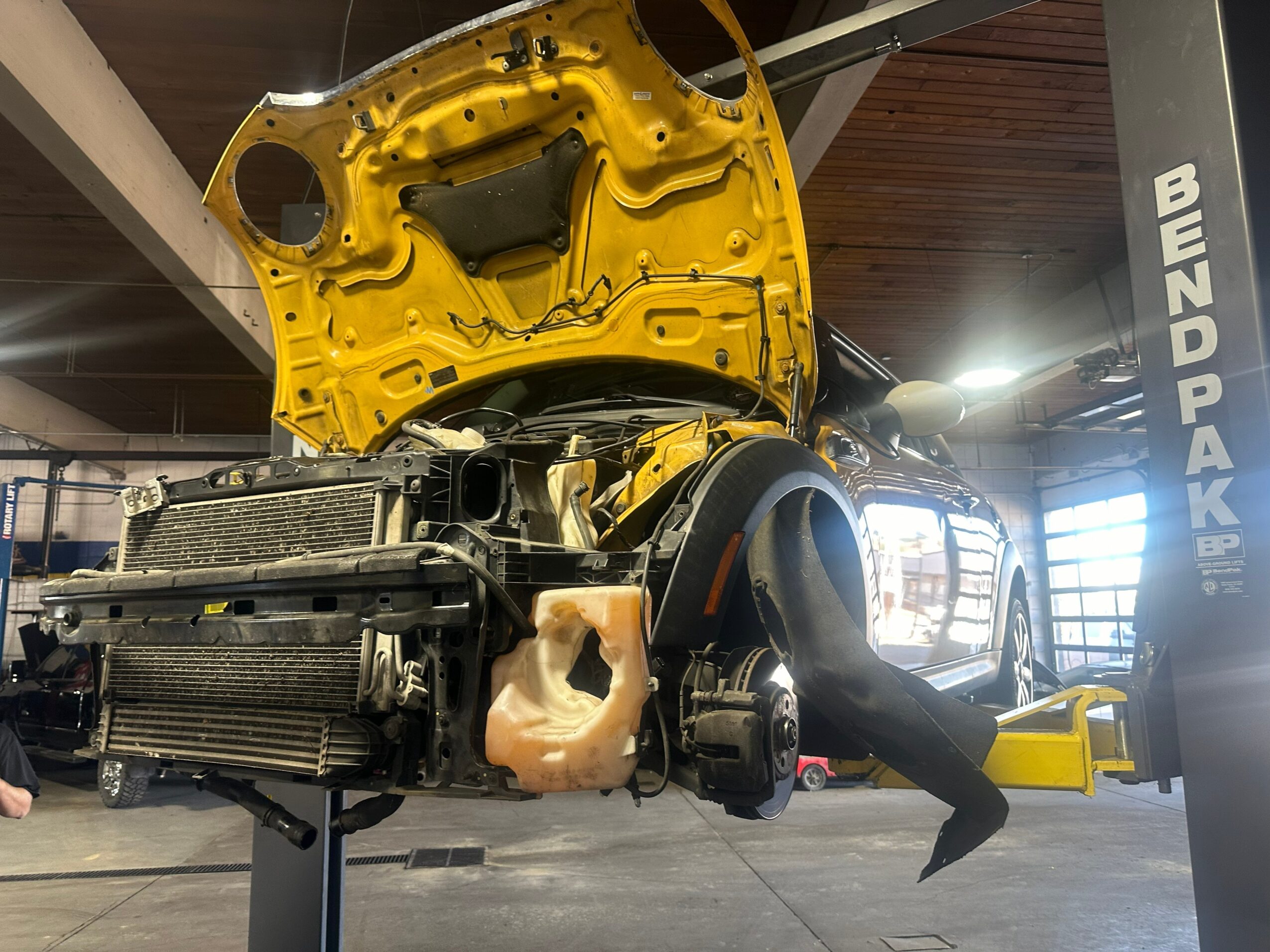 Car with open hood and front bumper removed, in a workshop, with a person working underneath, and a yellow lift nearby.