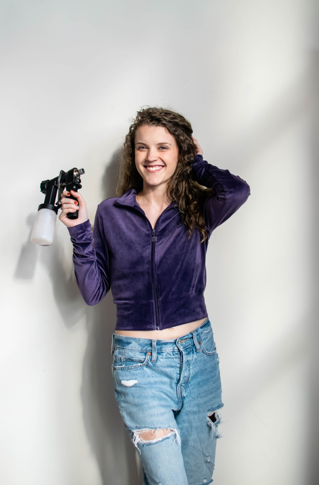Young woman with curly hair smiling, holding a camera, wearing a purple jacket and ripped jeans, standing against a plain background.