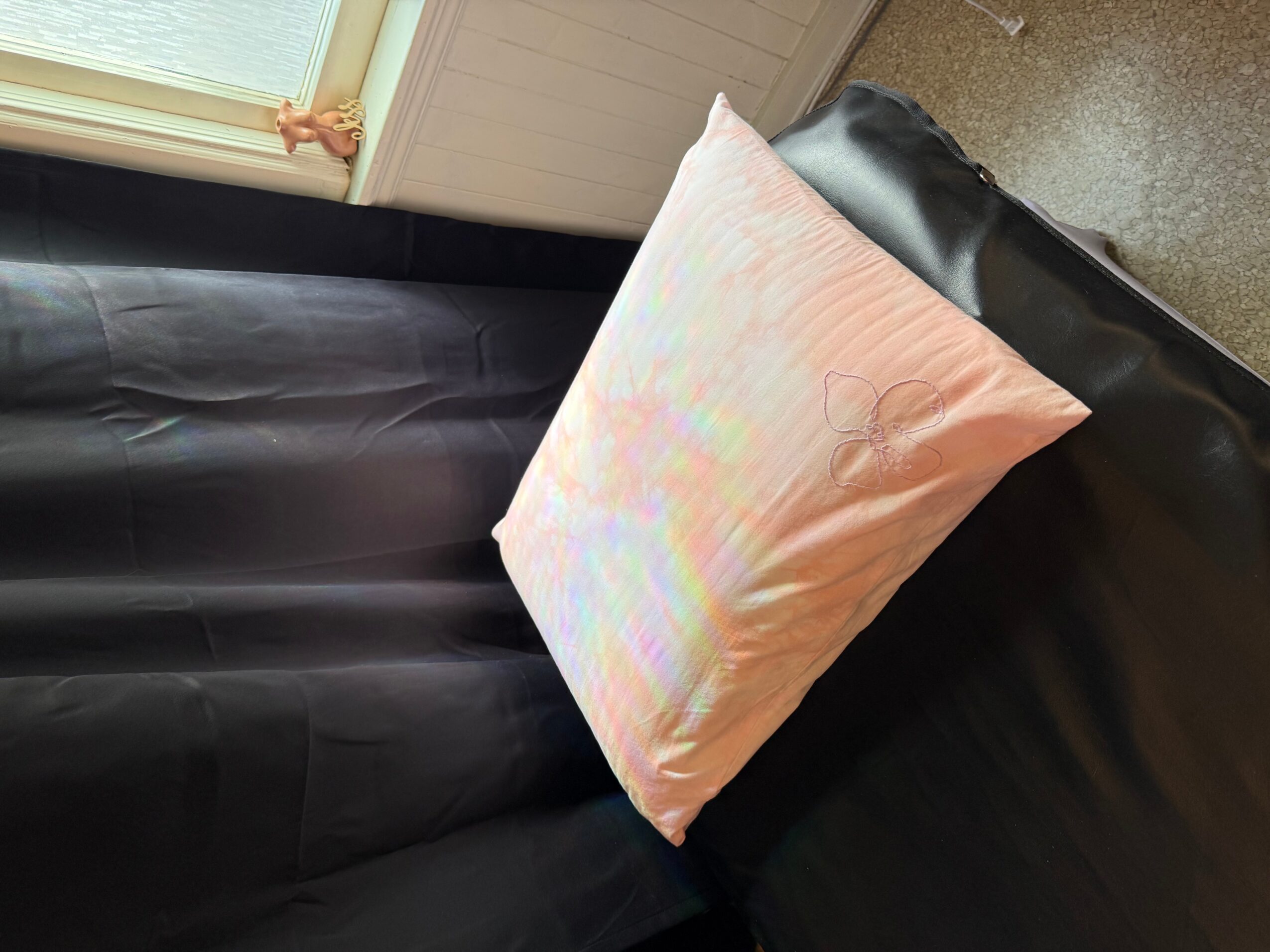 Pink pillow with a subtle pattern resting on a black surface, near a window with a white frame.
