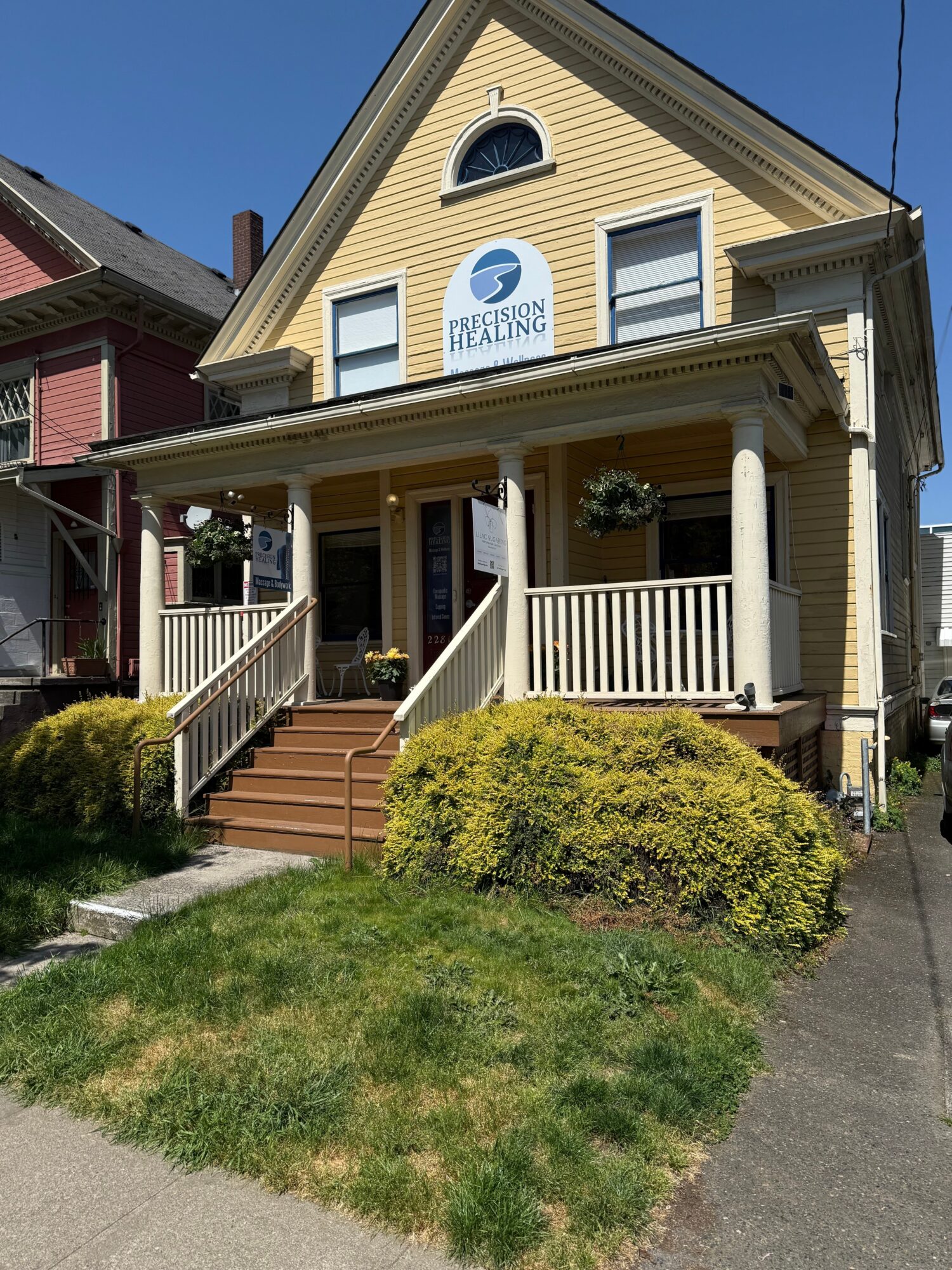 Yellow house with a front porch, stairs, and hanging flower baskets, under a clear blue sky.