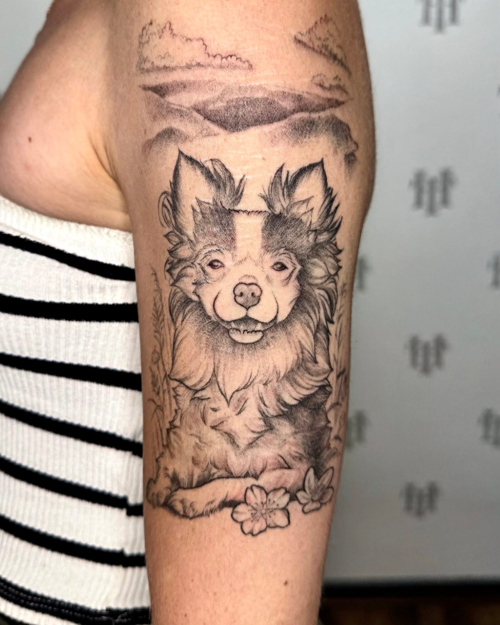 Black and gray tattoo of a smiling dog with a flower, on person's upper arm, with clouds in background.