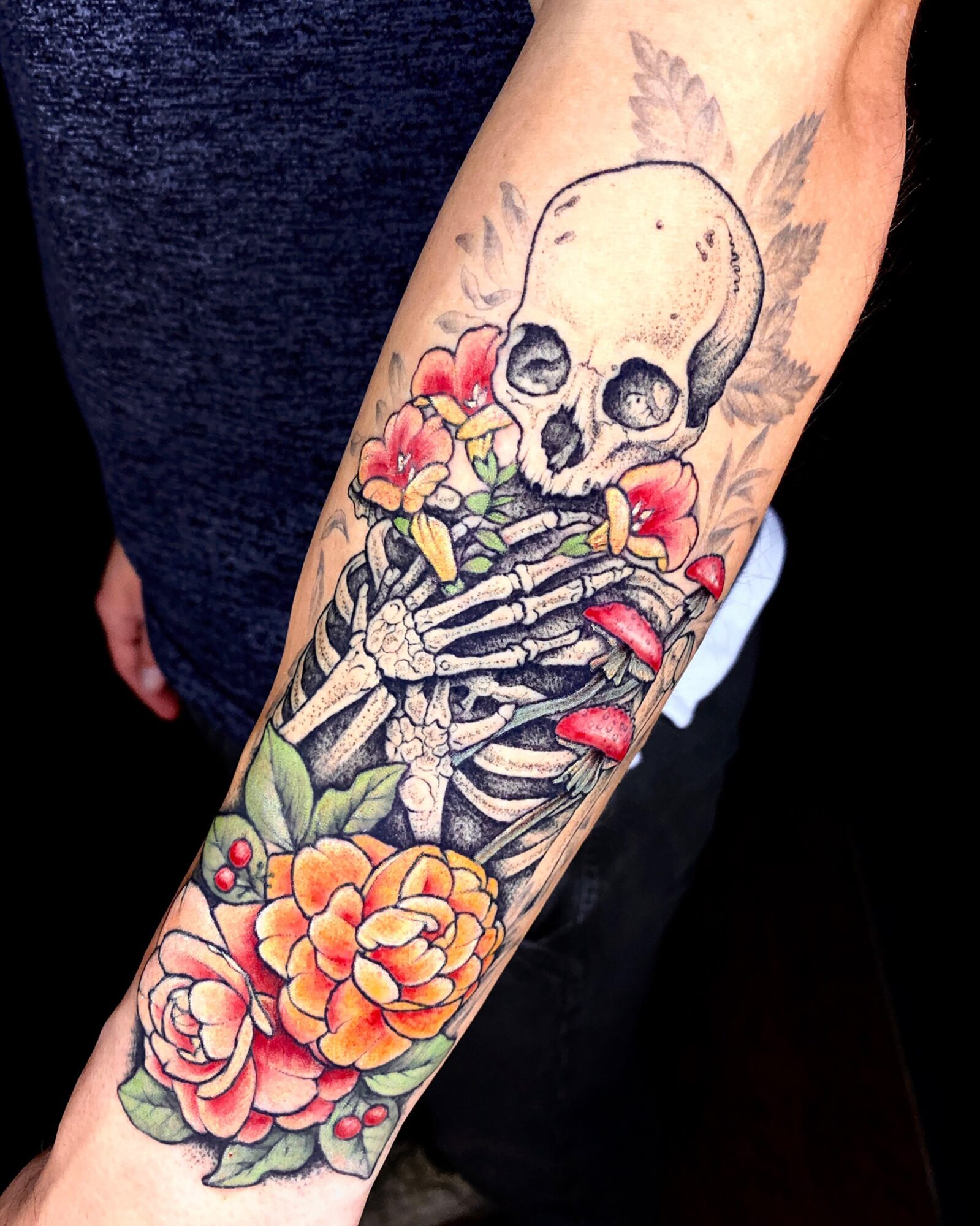 Tattoo of a skull with flowers and a skeleton hand on a person's arm, colorful and detailed.