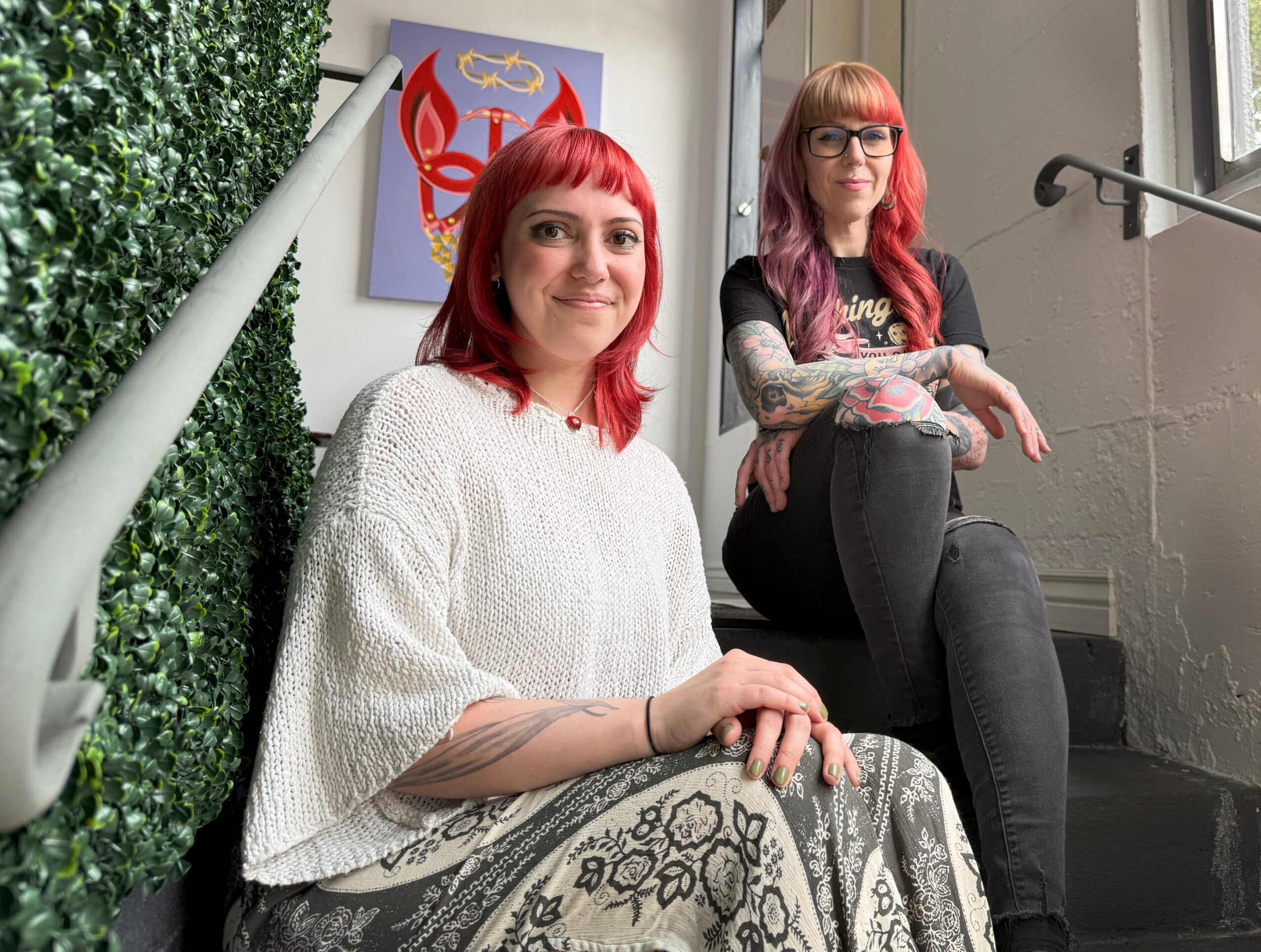 Two women with colorful hair sitting on stairs indoors, one smiling at camera, the other looking away, with artwork on wall behind them.