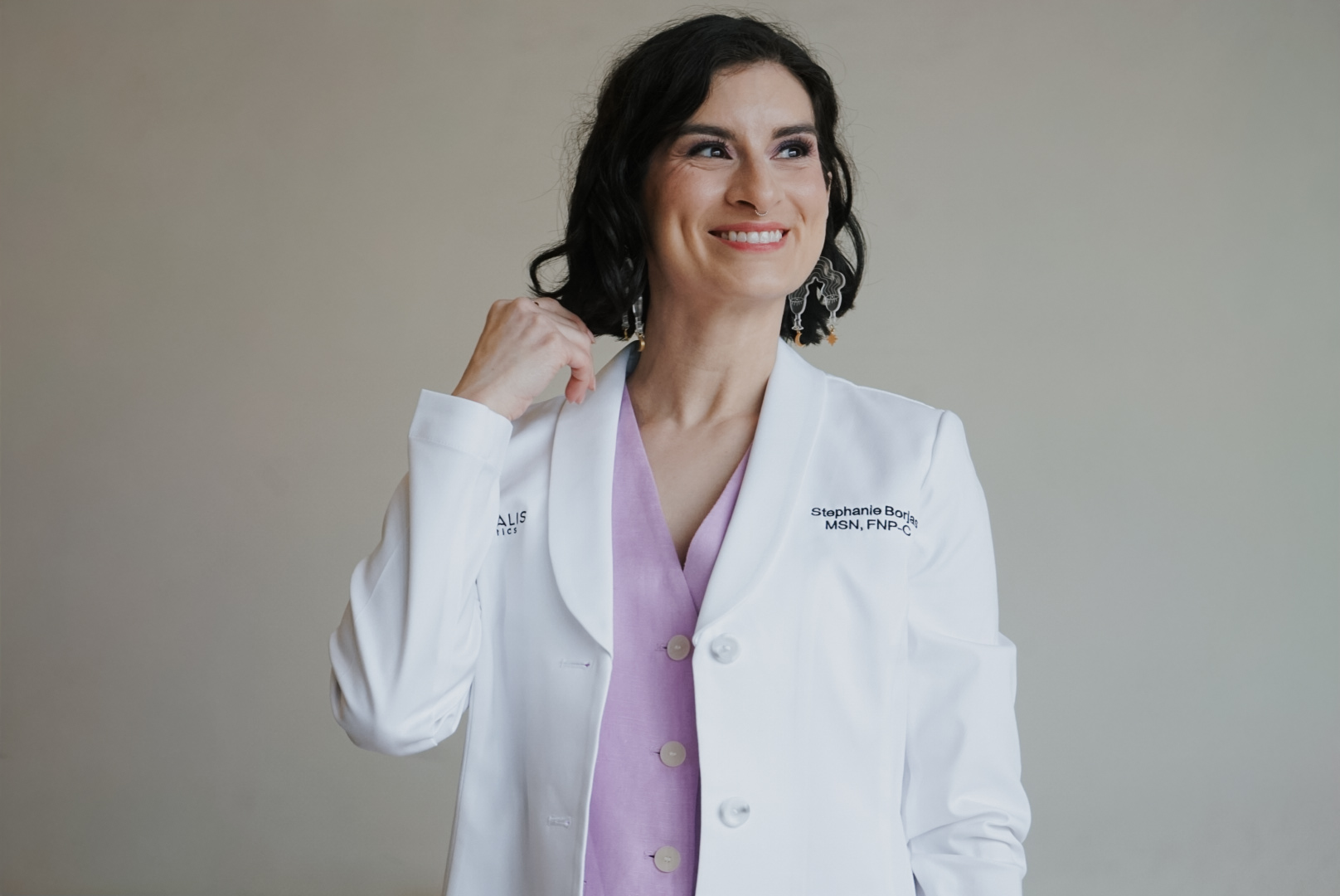 Woman in white medical coat smiling, wearing purple shirt, against plain background.