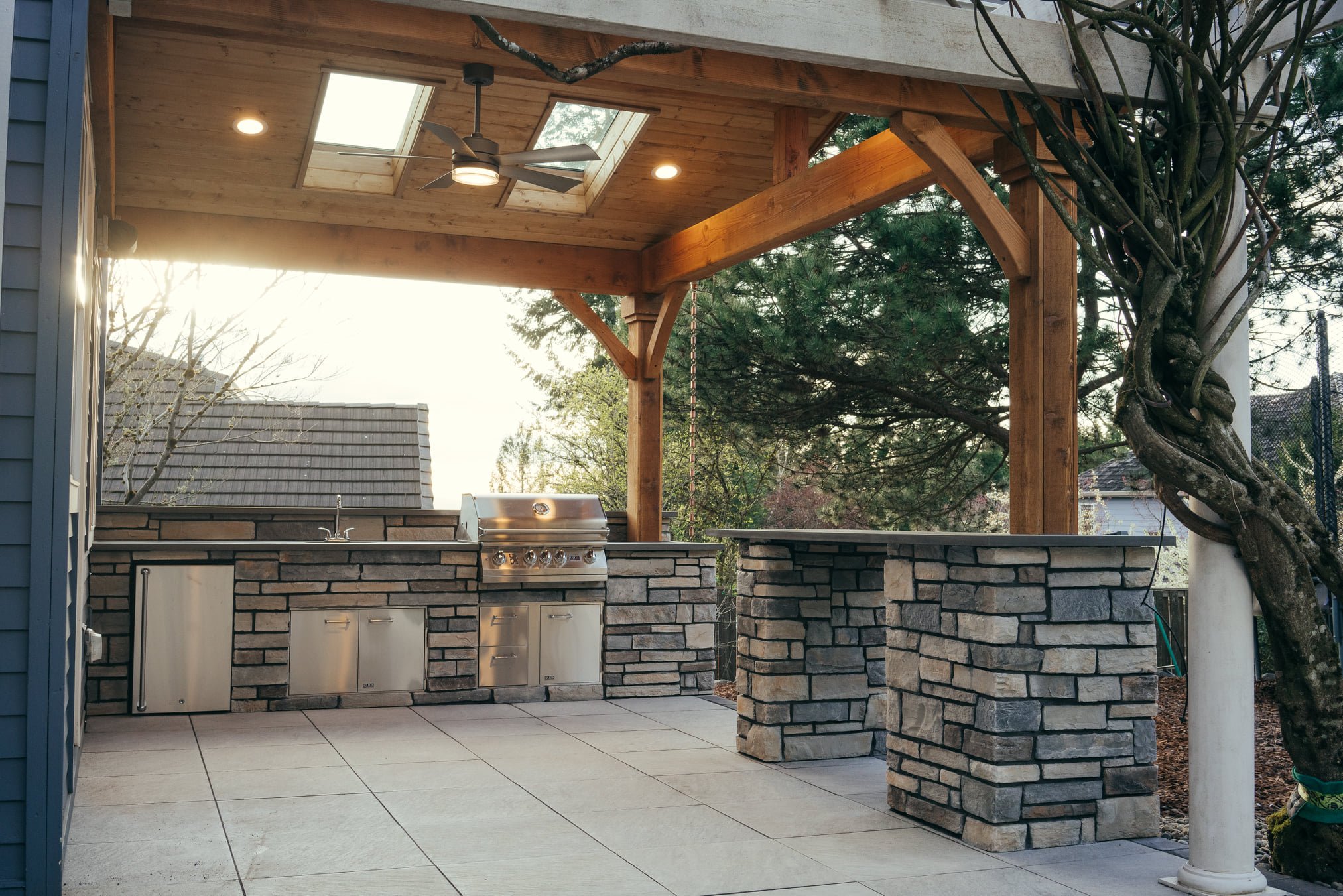 Outdoor kitchen with stone counters, built-in grills, and a wooden pergola, surrounded by trees and neighboring houses.