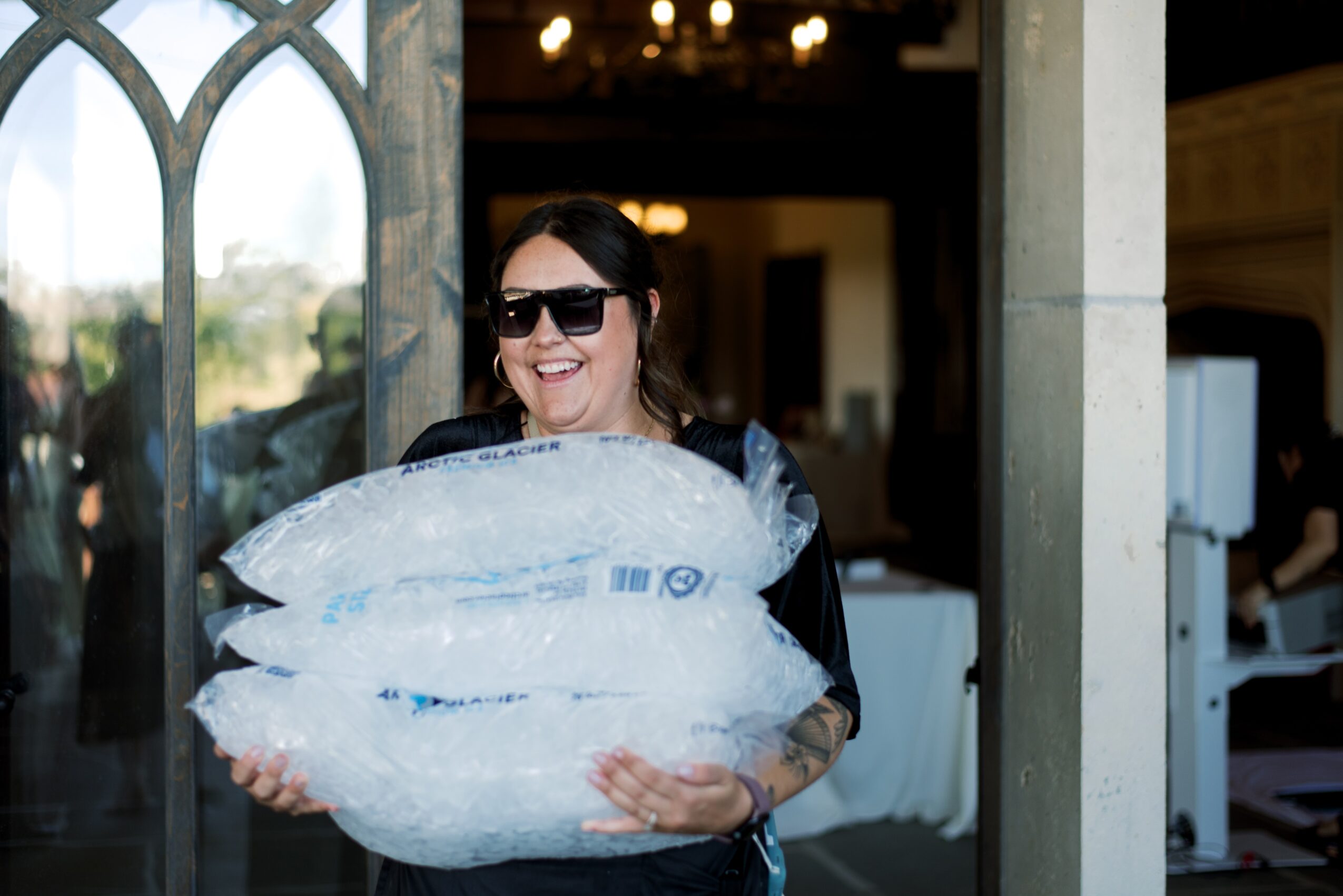 Woman wearing sunglasses holding a large fish, smiling indoors with arched window and chandelier in background.